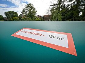 Eine große, grüne, faltbare Zisterne, die eingezäunt auf einer Wiese aufgestellt ist. Im Hintergrund sind zwei MIDEWA-Transporter. Die Zisterne ist mit Aufklebern versehen, das auf die Nutzung für Löschwasser hinweist. Zisternen sind Behälter, in denen Wasser gespeichert wird.
