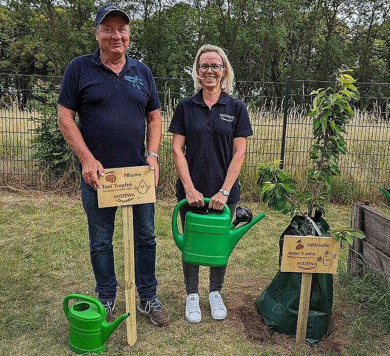 Ein Mitarbeiter und eine Mitarbeiterin der MIDEWA stehen in einem Schulgarten. Anlass ein Schulgartenprojekt was MIDEWA unterstützt. Beide stehen neben einem jungen frisch eingepfanztem Kirschbaum mit Bewässerungssack. Der Mann im blauen MIDEWA-Shirt hält ein Holzschlild und die Frau hält eine grüne Giesskanne in der Hand. 