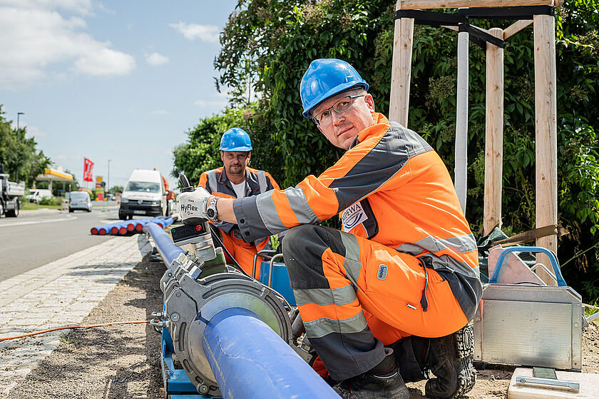Zwei MIDEWA-Mitarbeiter in orangefarbener Kleidung hocken vor Trinkwasserrohren und reparieren eine defekte Leitung. Beide tragen blaue Helme. 
