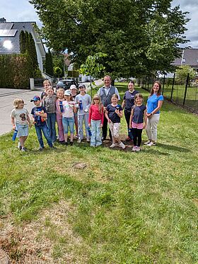 MIDEWA-Mitarbeiter, der Bürgermeister Martin Blümel und die Hortkinder der Grundschule in Röblingen stehen an der gepflanzten Winterlinde. Im Hintergrund sind gründe Bäume und ein grüner Rasen. Es ist ein sonniger Tag.