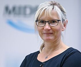 Anja Stollberg, Leiterin im Kundenservice der MIDEWA steht vor einem Büro. Sie trägt eine schwarze Bluse.