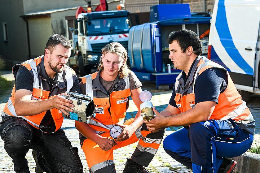 Drei junge Männer in orangfarbender Arbeitskleidung knien nebeneinander auf einem gepflastertem Weg. Alle Männer halten typische Materialien der Wasserwirtschaft in der Hand. Der Mann in der Mitte ist ein Auszubildender und bekommt von seinen Lehrmeistern eine Wasseruhr erklärt. Im Hintergrund sind zwei MIDEWA Fahrzeuge zu sehen.