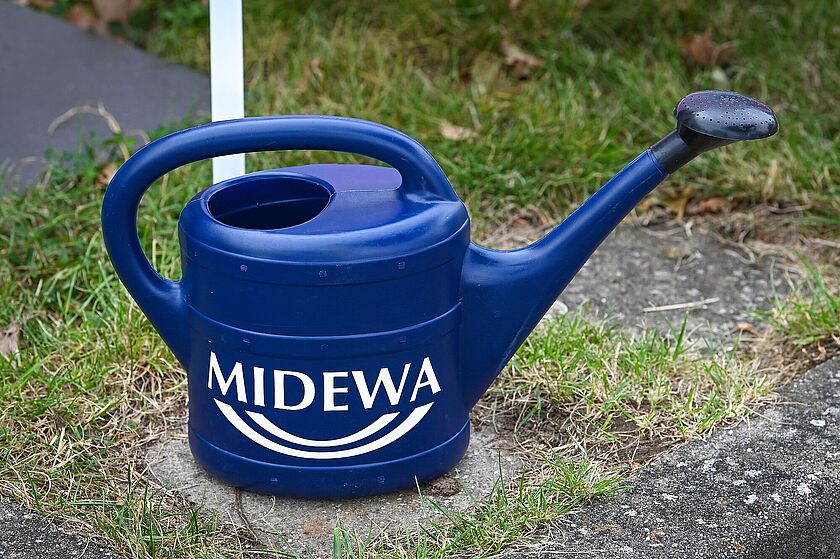 Eine blaue Gießkanne mit MIDEWA Logo steht auf einer Rasenfläche.