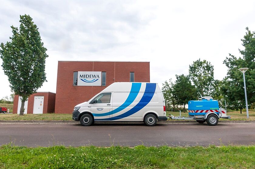Ein MIDEWA-Transporter beklebt mit zwei blauen Wellen und einem blauen Wasserwagenanhänger steht vor einem  Gebäude. Auf dem rot gepflastertem Gebäude ist eine großes Schild mit dem Firmenlogo der MIDEWA angebracht. 