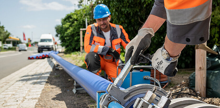 Das Bild zeigt den Ausschnitt einer Baustelle. Ein Trinkwassermonteur in orangener Arbeitskleidung und blauem Schutzhelm kniet an einer Trinkwasserleitung. Im Vordergrund sieht man Hände eines zweiten Trinkwassermonteurs. Dieser trägt Handschuhe und bearbeitet mit Spezialwerkzeug das Trinkwasserrohr. 