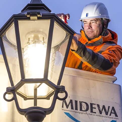 Ein MIDEWA-Mitarbeiter in orangener Arbeitskleidung führt Wartungsarbeiten an einer historischen Straßenlaterne durch. Er trägt einen weißen Arbeitshelm und nutzt für die Arbeiten eine Hubsteigerbühne. Ein Ausschnitt des weißen Arbeitskorbes ist zu sehen.