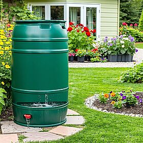 Eine grüne Regentonne steht auf einer Steinfläche in einem Garten. Im Hintergrund sind Beete mit bunten Blumen sowie eine Gartenlaube zu sehen.